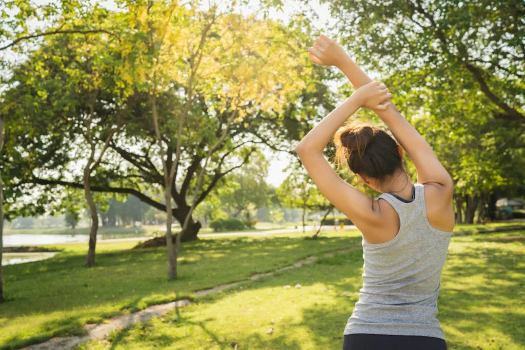 Mulher de regata alongando os braços em um parque arborizado, vista de costas
