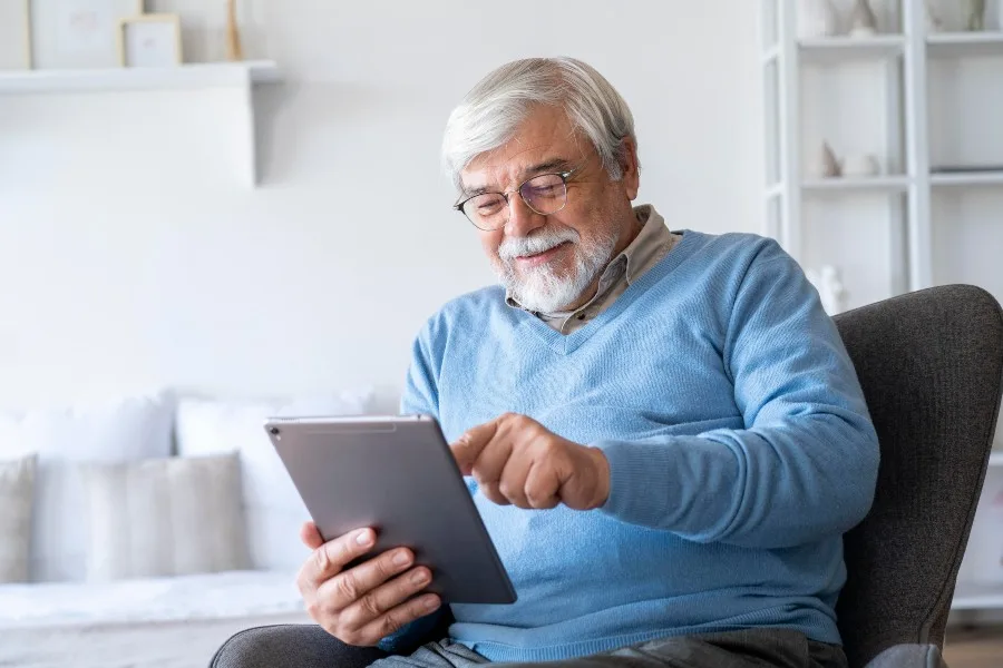 Homem idoso sorrindo enquanto usa um tablet sentado em poltrona na sala de casa