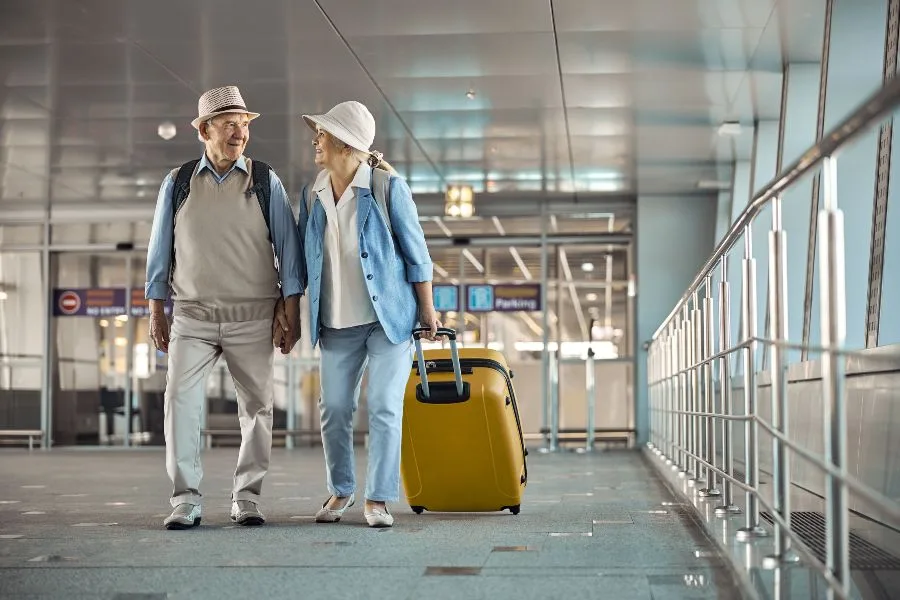 Casal de idosos caminhando de mãos dadas com mala amarela no corredor de um aeroporto