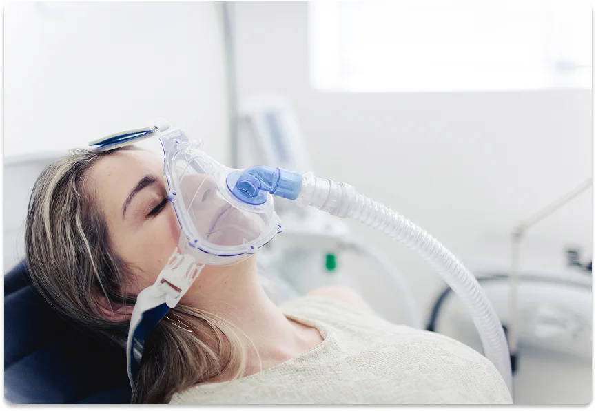 Paciente deitada com máscara de sedação no rosto e olhos fechados, em sala de procedimento