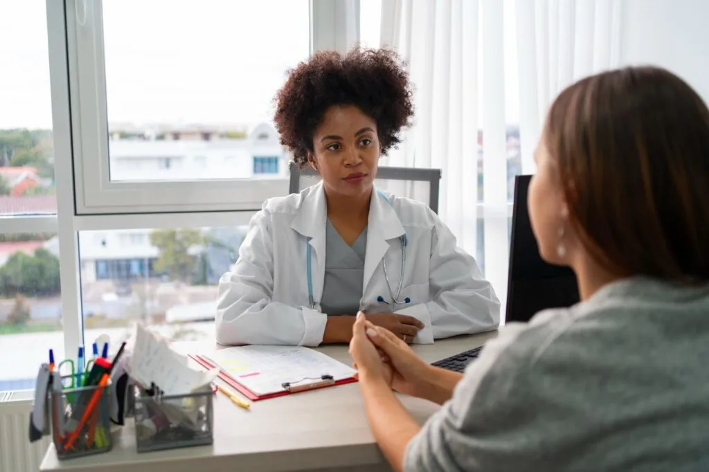 Médica conversando com uma paciente sentada à mesa do consultório