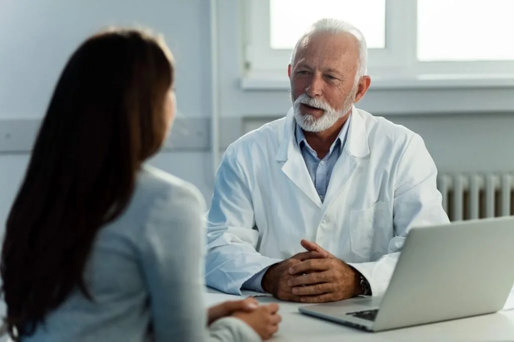 Médico de jaleco conversando com uma paciente à mesa do consultório