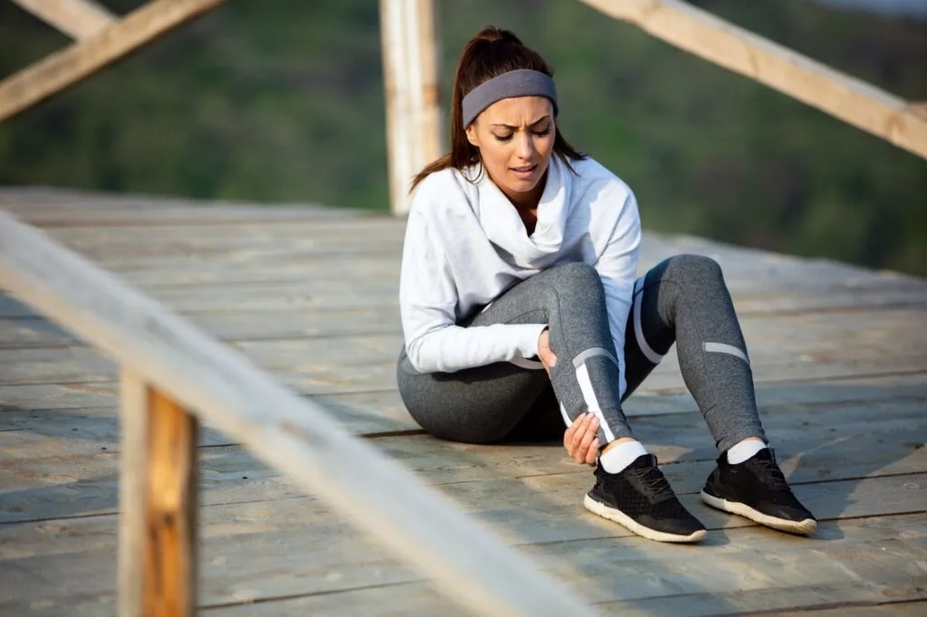Mulher em roupas de corrida sentada em um deck de madeira segurando a panturrilha com dor