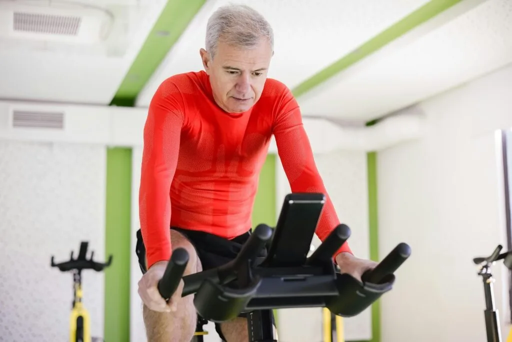 Homem grisalho de camiseta vermelha pedalando em bicicleta ergométrica na academia