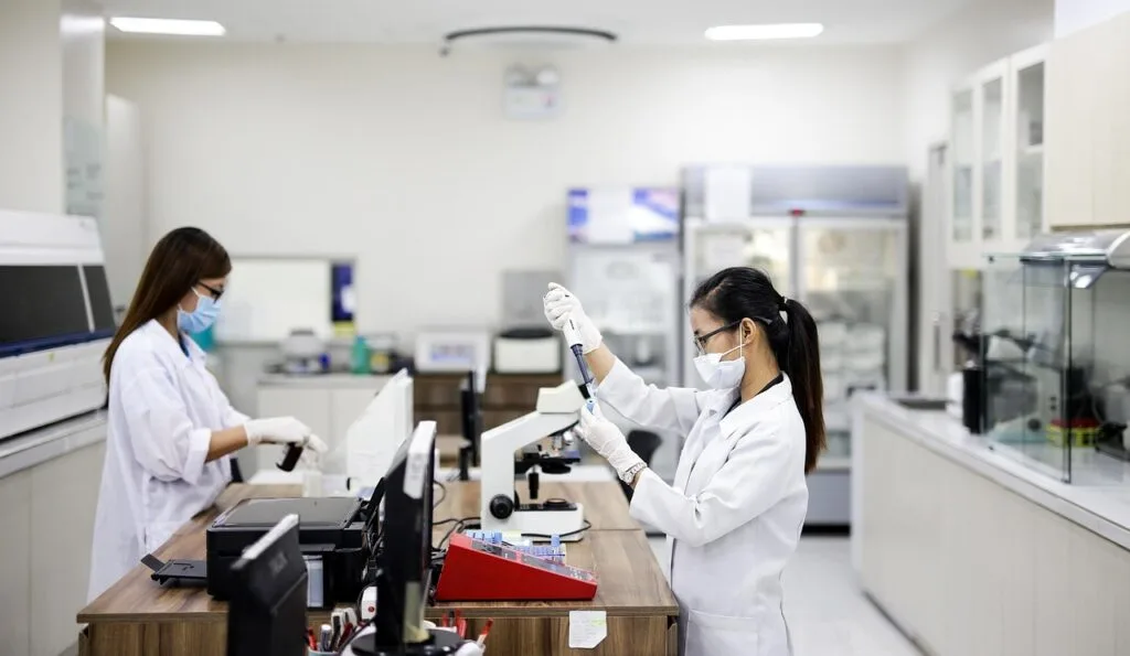 Duas profissionais de jaleco e máscara trabalham em bancada de laboratório de análises clínicas