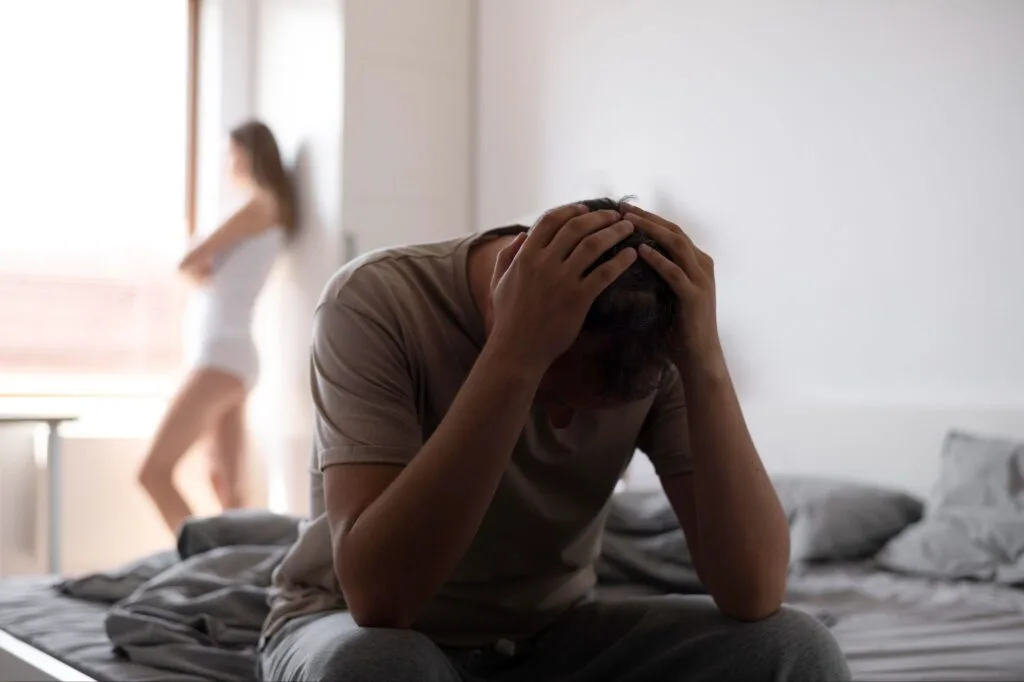 Homem sentado na beira da cama com as mãos na cabeça e uma mulher desfocada ao fundo