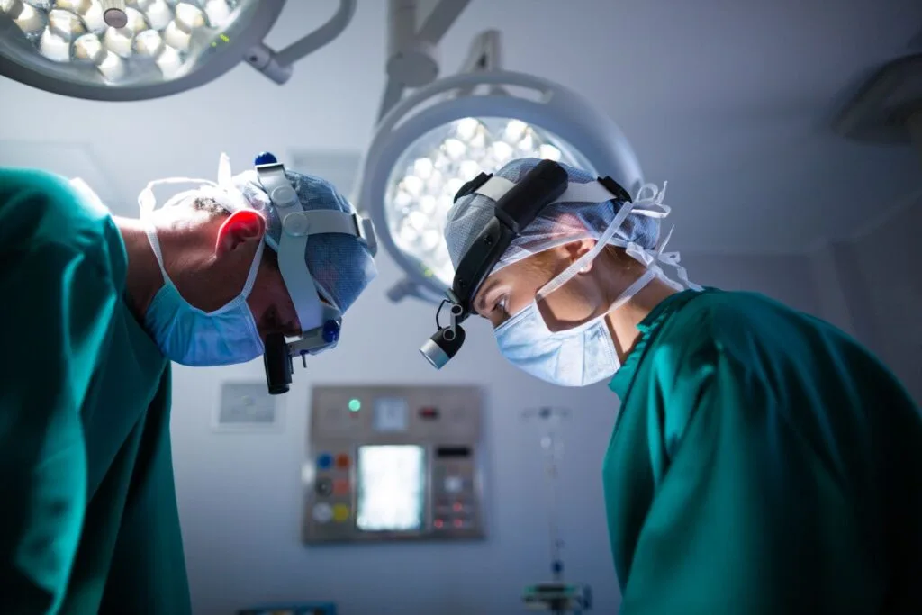 Dois cirurgiões de máscara e lupa cirúrgica inclinados sob os focos de luz da sala de cirurgia