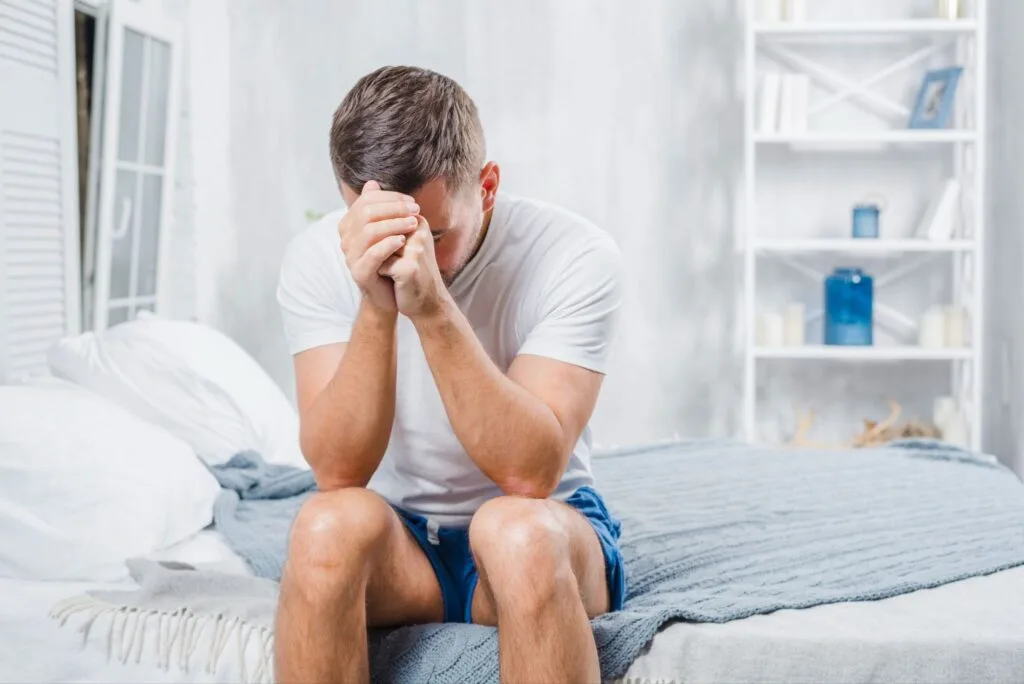 Homem de camiseta branca sentado na cama, cabisbaixo, com a testa apoiada nas mãos