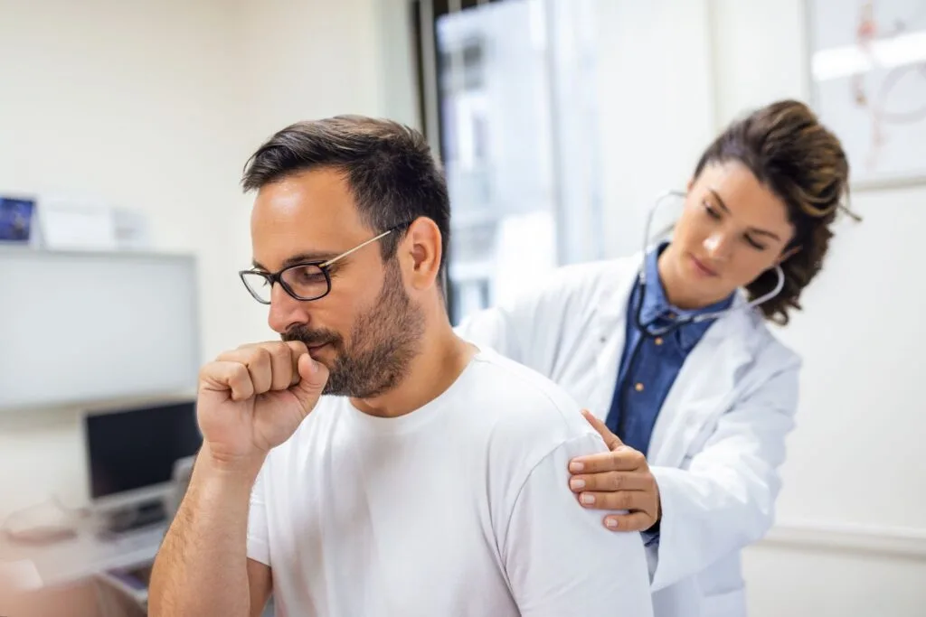 Homem tosse com a mão na boca enquanto médica o ausculta pelas costas no consultório