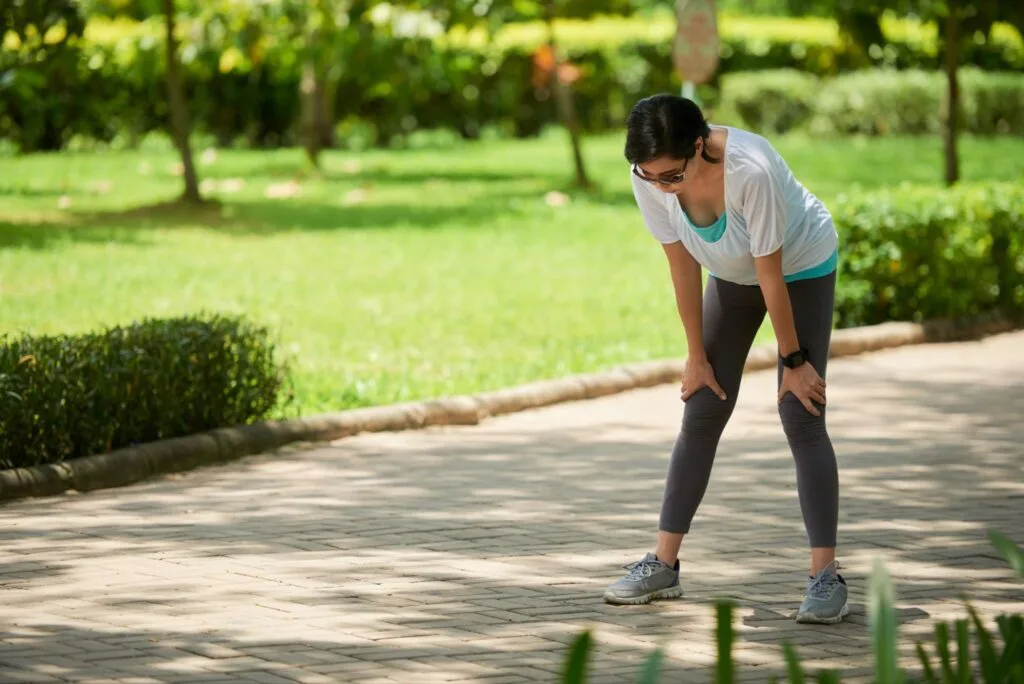 Mulher curvada com as mãos nos joelhos fazendo pausa durante caminhada em um parque