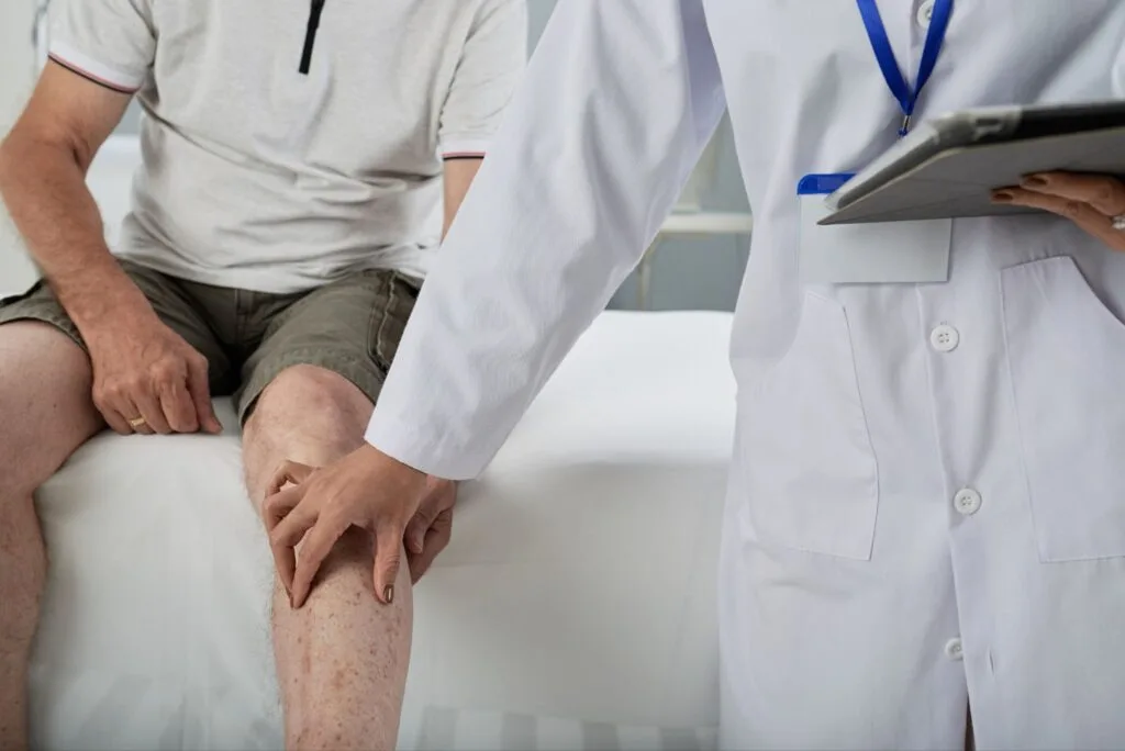 Profissional de jaleco examina a perna de um paciente sentado na maca do consultório